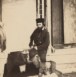 Black and white photograph of a bearded man sitting on a bench in front of a wooden building.